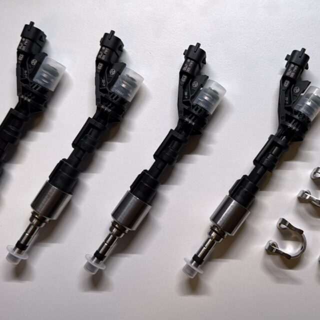 Four automotive fuel injectors arranged diagonally on a light surface, with metal clamps placed nearby as tools for installation or maintenance.